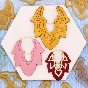 May include: Three pairs of large, decorative hoop earrings with intricate cut-out designs. The earrings are made of wood and come in gold, pink, and burgundy.