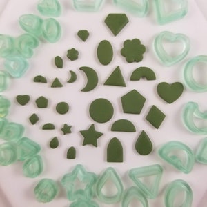 May include: A collection of green polymer clay shapes, including hearts, stars, moons, and geometric shapes. The shapes are arranged on a white surface.