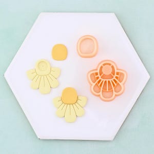 May include: A collection of flower-shaped cookie cutters and cut-out cookies in shades of yellow and orange. The cutters are made of plastic and the cookies are on a white hexagonal tile.