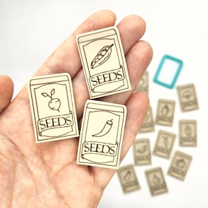 May include: Three wooden seed packets with illustrations of a pea pod, a radish, and a chili pepper. Each packet has the word "SEEDS" printed on it.