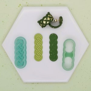May include: A set of five silicone molds for making jewelry. The molds are in the shape of a braid, a circle, a wavy line, and two different shapes with holes. The molds are on a white surface.