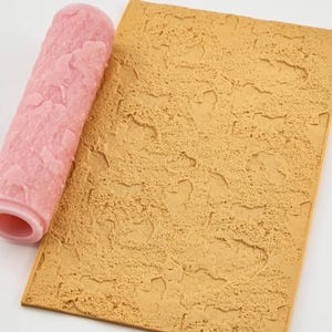 May include: A pink textured paint roller and a tan textured rectangular surface. The roller has a rough, uneven pattern. The surface has a similar texture, with irregular shapes and a mottled appearance. The items are likely used for decorative painting or texturing.