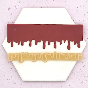 May include: Two silicone molds for baking, one red and one beige. The molds are shaped like strips with a dripping effect, resembling melted chocolate or caramel.