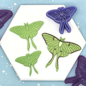 May include: Four green and purple butterfly cookie cutters on a white hexagonal surface. The cookie cutters are in various sizes and shapes.