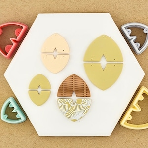 May include: A collection of polymer clay earring components in various colors and patterns. The components include oval shapes in beige, olive green, and a woven brown design with white floral accents. Several earring cutters in red, teal, gray, and yellow surround the components.