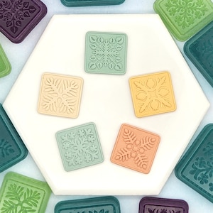 May include: A collection of square, colorful decorative tiles with floral designs. The tiles are in shades of green, peach, yellow, and beige, arranged on a white hexagonal surface. Other tiles in shades of green and purple surround the central arrangement.