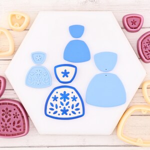 May include: A collection of blue and white cookie cutters in various shapes, including floral and abstract designs. The cutters are arranged on a white hexagonal surface, with additional cutters in pink and yellow.