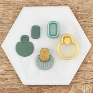 May include: A collection of polymer clay earring cutters in various shapes and colors. The cutters include a green rounded shape, a rectangle, an oval, and a yellow and white rounded shape. The cutters are arranged on a white hexagonal tile.