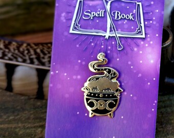 May include: Gold enamel pin featuring a black cauldron with a crescent moon, sun, and star design. The cauldron is steaming and has a snake wrapped around it. The pin is on a purple card with the words "Spell Book" in white lettering.