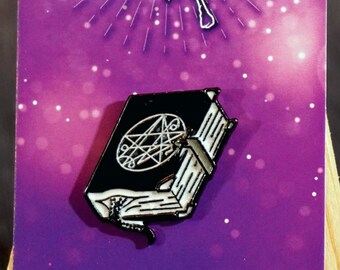 May include: A black enamel pin of a closed book with a pentagram on the cover. The pin is on a purple card with the words "Spell Book" and a stylized image of an open book with a wand.