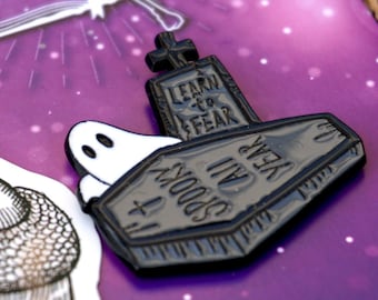 May include: A black enamel pin shaped like a coffin with the text "Learn to Fear" and "Spooky Jail" on it. The pin is on a purple background with a book and a ghost.