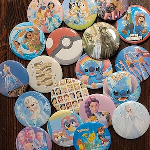 May include: A collection of colorful round button pins featuring various cartoon characters and pop culture references. Designs include Disney princesses, Bluey, Super Mario Bros, Pokemon, and Stitch. The pins are arranged on a dark wooden surface.