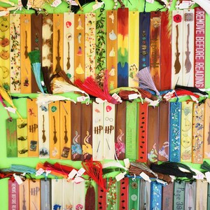 May include: A collection of colorful, handmade bookmarks with various designs, including images of a broom, a princess, and the text "REMOVE BEFORE READING." Each bookmark has a tassel. The background is a bright green.