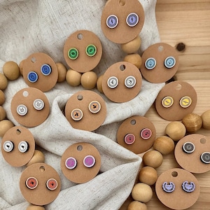 May include: A collection of colorful stud earrings with various designs, including symbols and icons, displayed on small, round cardboard tags. The earrings are in shades of blue, green, orange, pink, and purple, with wooden beads scattered around.