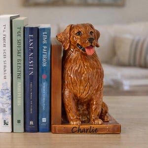 Peut inclure: Un serre-livres en céramique brune représentant un golden retriever avec la langue sortie, assis sur une base rectangulaire avec le nom Charlie imprimé en écriture noire, placé à côté d'une rangée de quatre livres reliés sur une surface plane.
