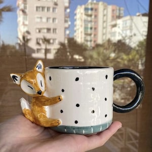 Könnte beinhalten: Eine weiße Keramiktasse mit schwarzen Punkten und einem blauen und grünen Streifen am Boden. Eine braune und orangefarbene Keramikfuchsfigur ist an der Seite der Tasse befestigt.