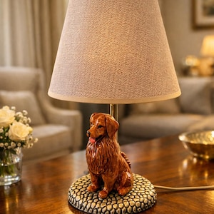May include: A decorative table lamp featuring a brown dog figurine base. The lamp has a beige fabric shade and a gold-colored metal stem. The dog figurine is detailed with a textured fur appearance and a red tongue. The lamp sits on a wooden table.