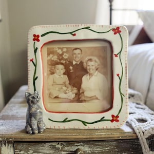 May include: A white ceramic picture frame with a family photo, decorated with green vines and red flowers. A gray cat figurine sits on a distressed wooden table next to the frame. The photo shows a family of three.