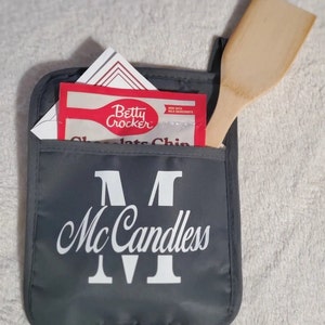 May include: Gray oven mitt with a white embroidered monogram "M Mc Candless IV". The mitt has a pocket with a Betty Crocker chocolate chip cookie mix packet and a wooden spatula inside.