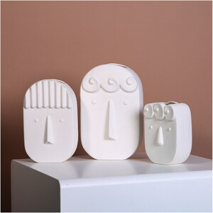 May include: Three white ceramic vases shaped like faces with different facial features. The vases are on a white surface.