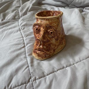 May include: A handmade ceramic vase with a face sculpted on the front. The vase is tan with brown accents, featuring a detailed face with open eyes and a defined mouth. The unique, artistic design adds a touch of character to any space.