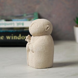 Puede incluir: Una peque&ntilde;a escultura de piedra blanca de una persona sosteniendo una flor. La escultura tiene un dise&ntilde;o simple y redondeado y una superficie texturizada.