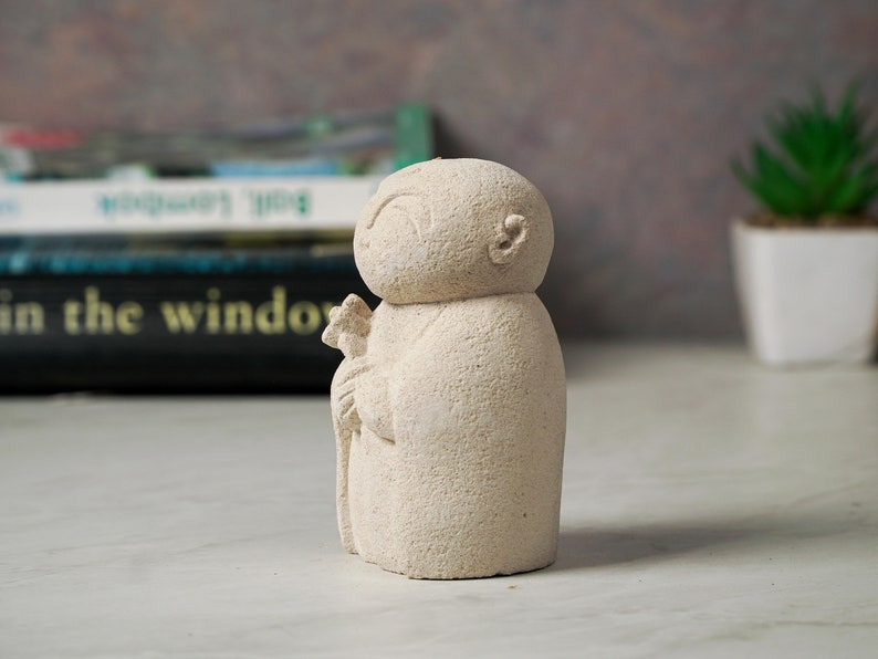 Puede incluir: Una peque&ntilde;a escultura de piedra blanca de una persona sosteniendo una flor. La escultura tiene un dise&ntilde;o simple y redondeado y una superficie texturizada.