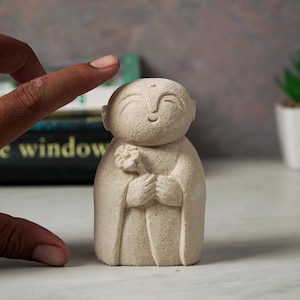 Puede incluir: Una peque&ntilde;a escultura de piedra blanca de un Buda sonriente sosteniendo una flor. La escultura est&aacute; hecha de un material rugoso y texturizado.
