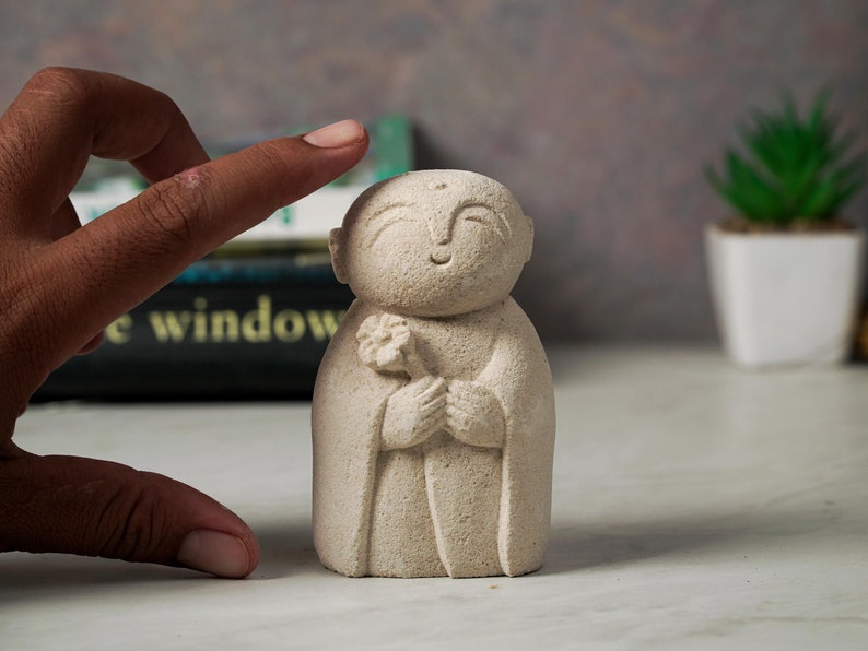 Puede incluir: Una peque&ntilde;a escultura de piedra blanca de un Buda sonriente sosteniendo una flor. La escultura est&aacute; hecha de un material rugoso y texturizado.