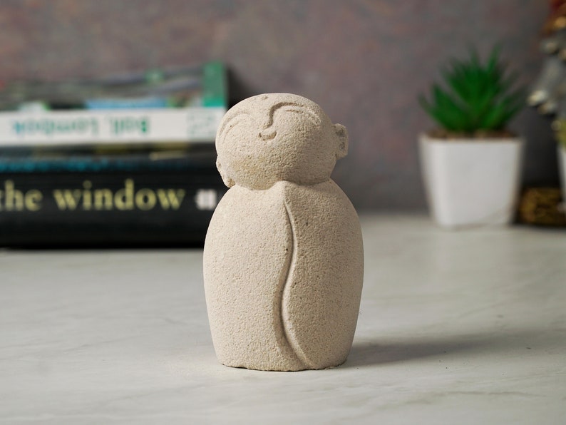 May include: A small, beige, stone sculpture of a person with their eyes closed. The sculpture has a smooth, textured surface and is shaped like a person with their arms crossed in front of them.