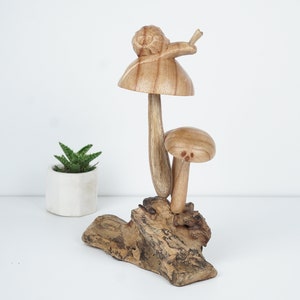 Puede incluir: Una escultura de madera de dos setas con un caracol en la parte superior de la seta m&aacute;s grande. Las setas son marrones y tienen una veta de madera natural. La escultura est&aacute; sentada sobre un trozo de madera a la deriva.