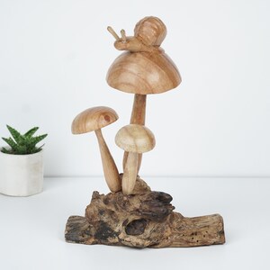 Puede incluir: Escultura de madera de tres setas con un caracol en la parte superior de la seta m&aacute;s grande. Las setas son marrones y blancas y est&aacute;n sobre un trozo de madera flotante.