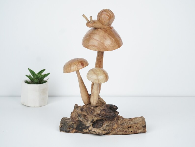 Puede incluir: Escultura de madera de tres setas con un caracol en la parte superior de la seta m&aacute;s grande. Las setas son marrones y blancas y est&aacute;n sobre un trozo de madera flotante.