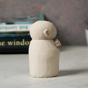 Puede incluir: Una peque&ntilde;a escultura de piedra blanca tallada a mano que representa a una persona con la cabeza redonda y un dise&ntilde;o simple y abstracto. La escultura est&aacute; sobre una superficie blanca.