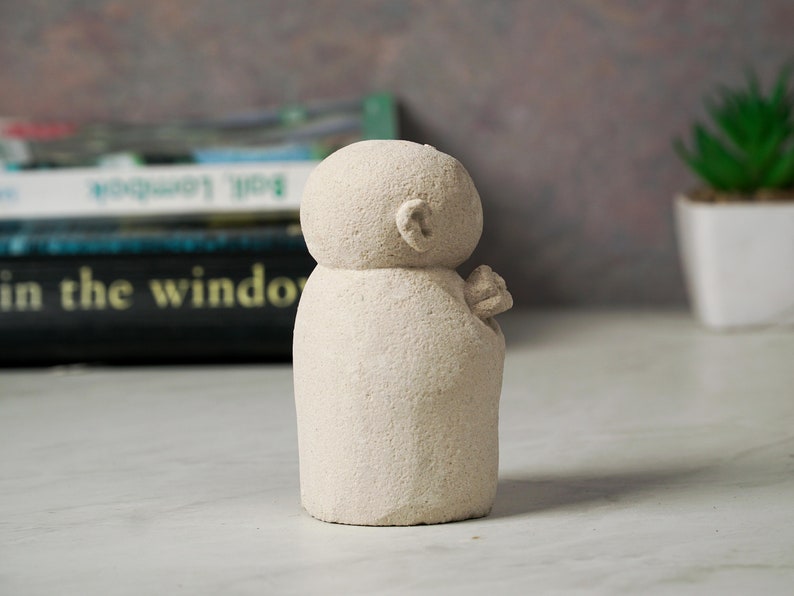 Puede incluir: Una peque&ntilde;a escultura de piedra blanca tallada a mano que representa a una persona con la cabeza redonda y un dise&ntilde;o simple y abstracto. La escultura est&aacute; sobre una superficie blanca.