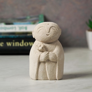 Puede incluir: Una peque&ntilde;a escultura de piedra blanca de una figura sonriente sosteniendo una flor. La escultura est&aacute; hecha de un material rugoso y texturizado y tiene un dise&ntilde;o simple y minimalista.