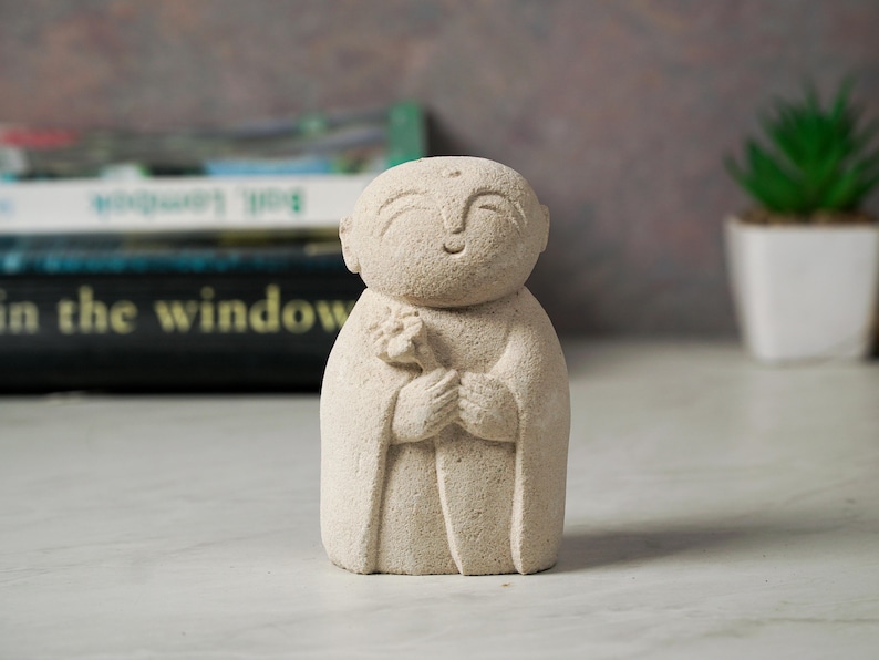 Puede incluir: Una peque&ntilde;a escultura de piedra blanca de una figura sonriente sosteniendo una flor. La escultura est&aacute; hecha de un material rugoso y texturizado y tiene un dise&ntilde;o simple y minimalista.
