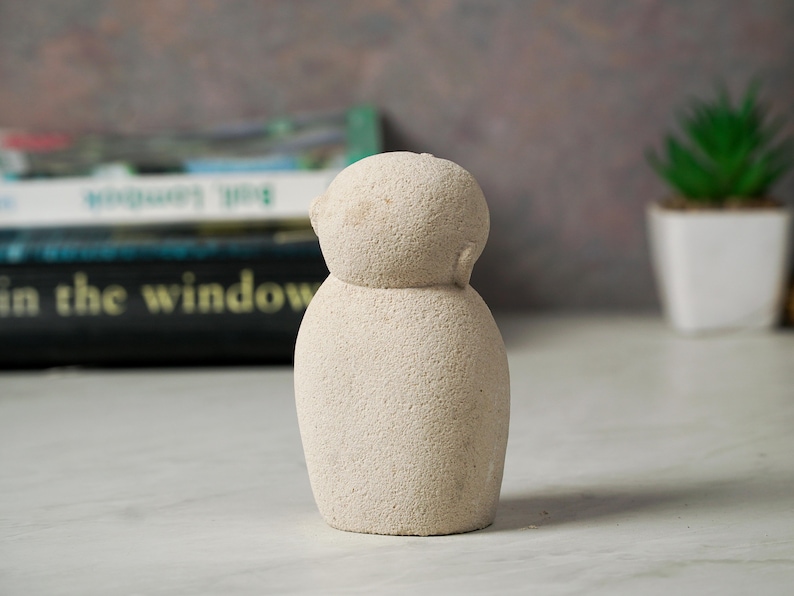 May include: A small, white, textured, ceramic sculpture of a person. The sculpture has a round head and a round body.