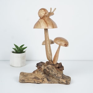 Puede incluir: Una escultura de madera de un hongo con un caracol en la parte superior, sentada sobre un trozo de madera a la deriva. El hongo es marr&oacute;n y tiene un tallo blanco. El caracol tambi&eacute;n es marr&oacute;n y tiene una concha blanca.