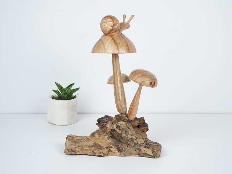 Puede incluir: Una escultura de madera de un hongo con un caracol en la parte superior, sentada sobre un trozo de madera a la deriva. El hongo es marr&oacute;n y tiene un tallo blanco. El caracol tambi&eacute;n es marr&oacute;n y tiene una concha blanca.