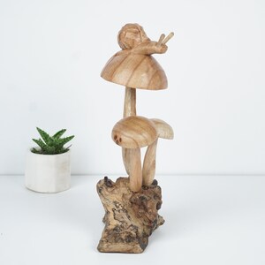 Puede incluir: Una escultura de madera de un caracol sentado encima de un hongo. El hongo est&aacute; sobre una base de madera. La escultura est&aacute; pintada en tonos marrones.