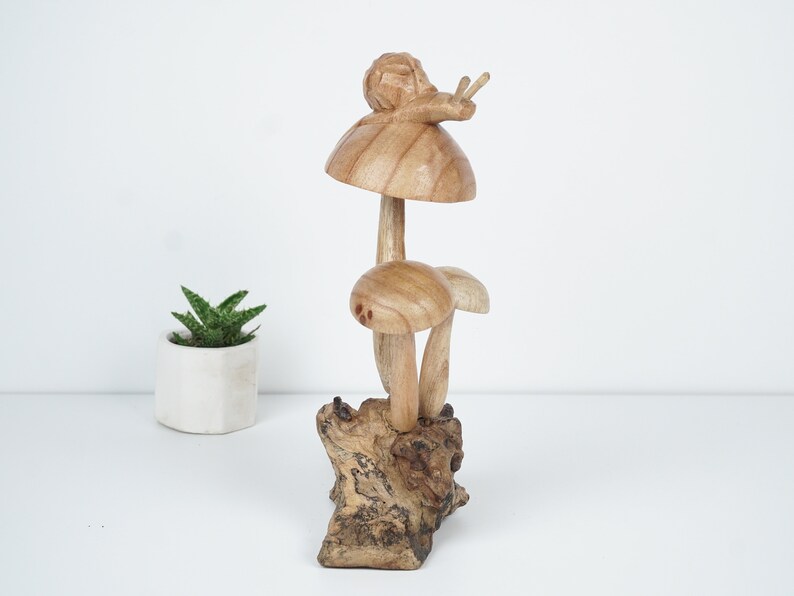 Puede incluir: Una escultura de madera de un caracol sentado encima de un hongo. El hongo est&aacute; sobre una base de madera. La escultura est&aacute; pintada en tonos marrones.