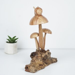 Puede incluir: Una escultura de madera de tres setas con un caracol en la parte superior, todo descansando sobre un trozo de madera a la deriva.