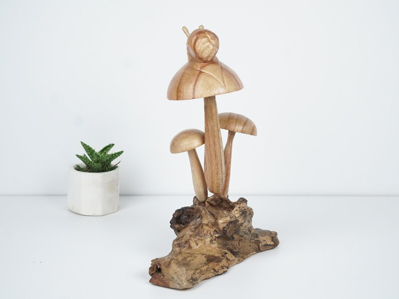 Puede incluir: Una escultura de madera de tres setas con un caracol en la parte superior, todo descansando sobre un trozo de madera a la deriva.