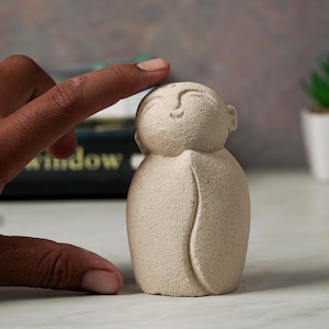 May include: A small, beige, hand-carved figurine of a smiling person with a rounded body and a simple design. The figurine is made of a textured material that resembles stone.