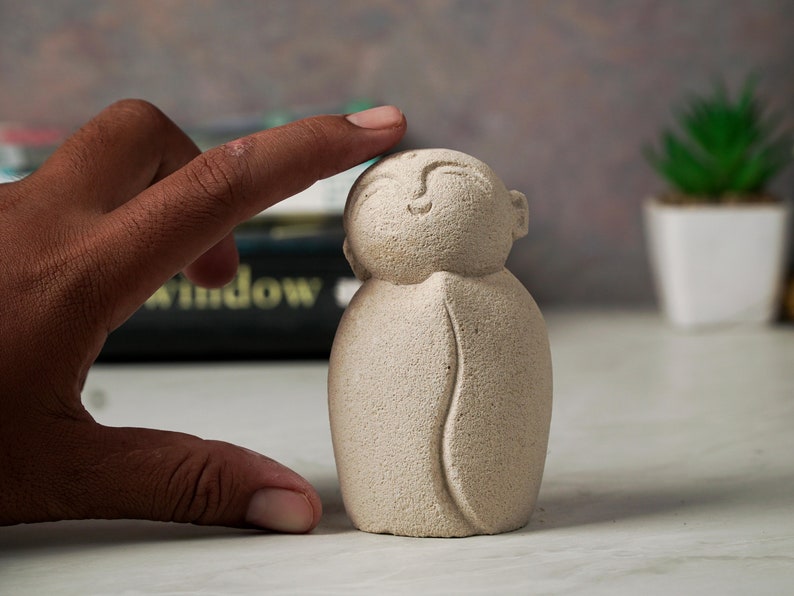 May include: A small, beige, hand-carved figurine of a smiling person with a rounded body and a simple design. The figurine is made of a textured material that resembles stone.