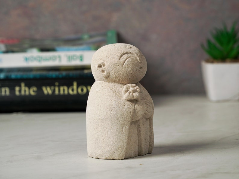 Puede incluir: Una peque&ntilde;a escultura de piedra blanca de una persona sosteniendo una flor. La escultura tiene una superficie texturizada y un dise&ntilde;o simple y minimalista.