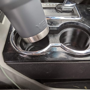 May include: A grey Yeti travel mug in a car cup holder. The cup holder is made of black plastic with a silver trim.