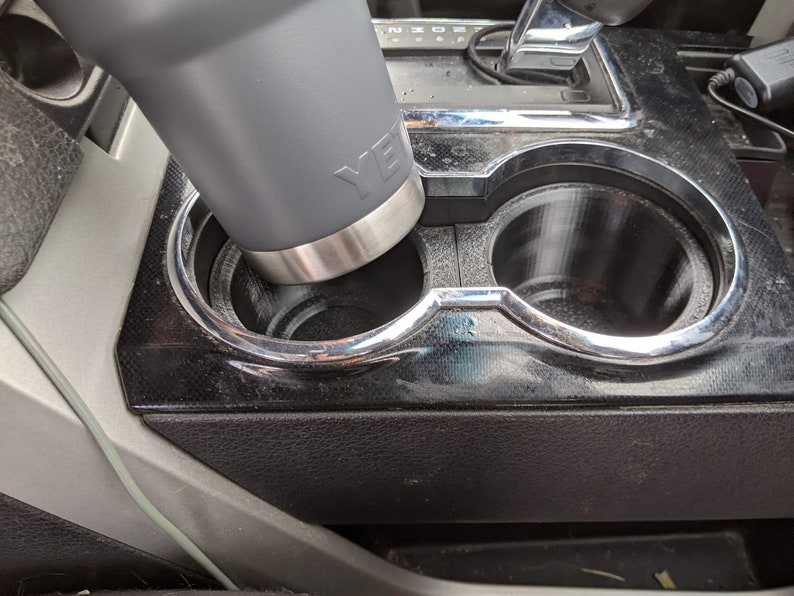 May include: A grey Yeti travel mug in a car cup holder. The cup holder is made of black plastic with a silver trim.