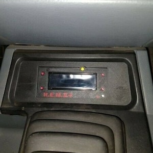 May include: Black dashboard component with a rectangular display and red text that reads "REBEL". The component has several indicator lights and is set in a gray interior.